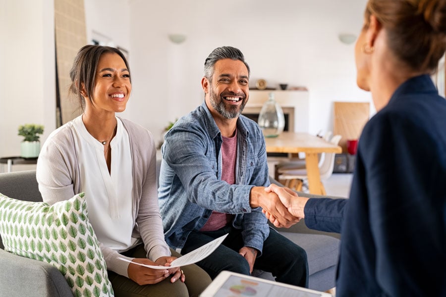 A couple sitting on a couch while the male is shaking the hand of a business person near the camera